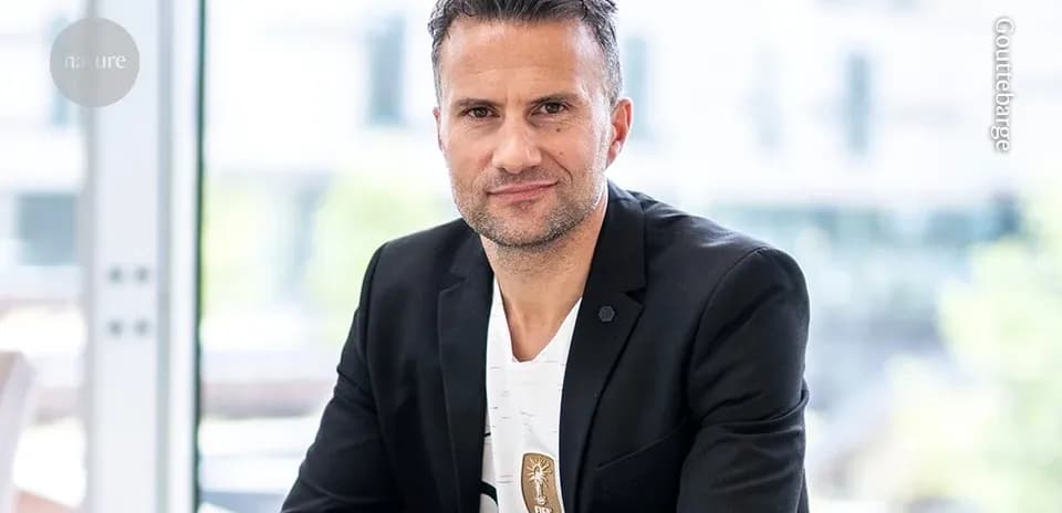 Man in black blazer seated indoors, looking at camera